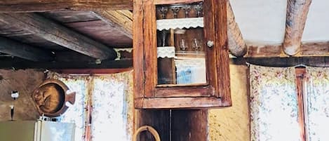 A rustic interior with a wooden ceiling, a small window with lace curtains, and a woven basket hanging on the wall.