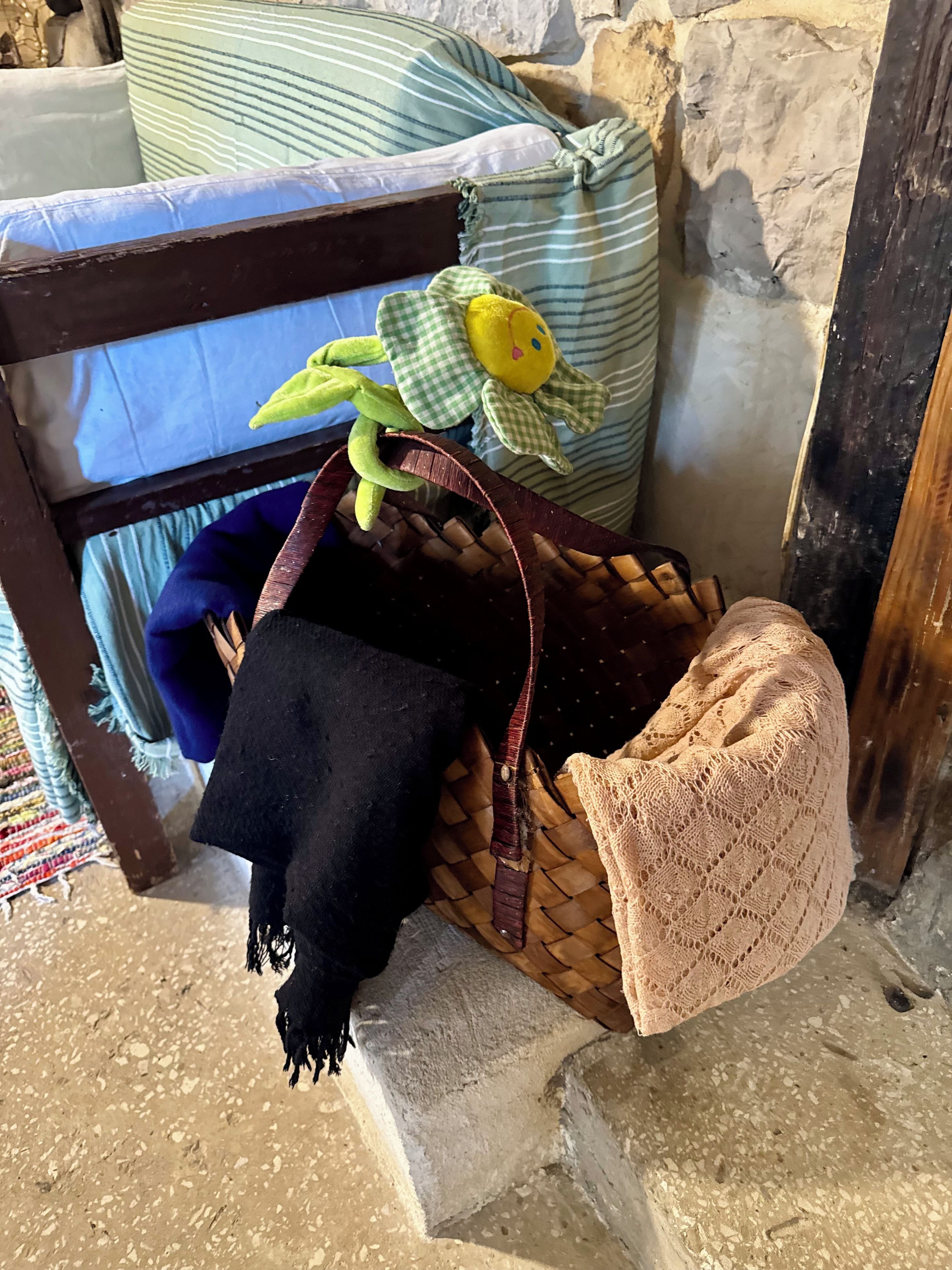 Una cesta de mimbre con un paño negro y un objeto tejido color beige, sobre un suelo de piedra junto a una cama con almohadones a rayas.