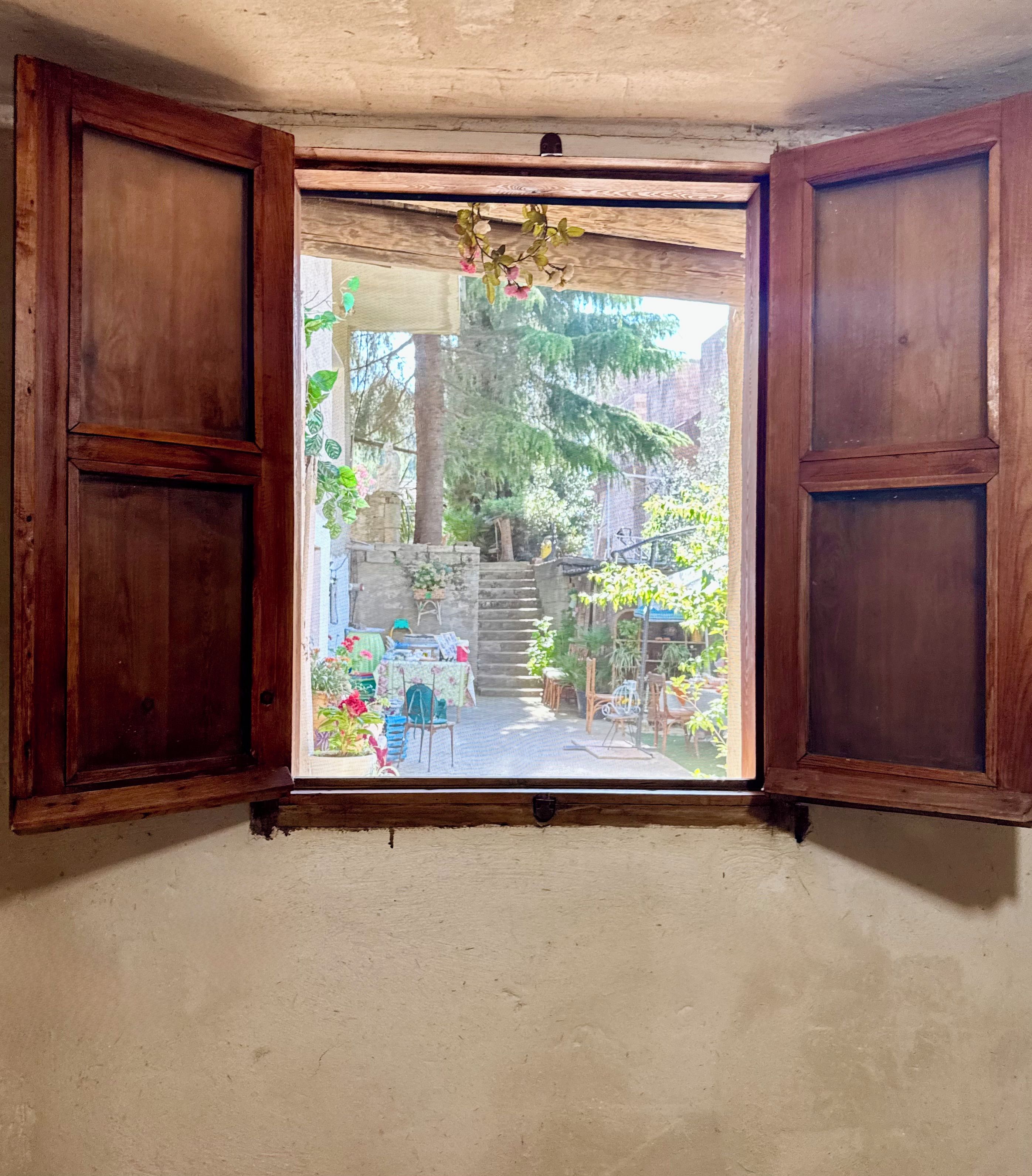 Een kamer met een raam met houten kader, met uitzicht op een tuin met planten en tuinmeubilair.