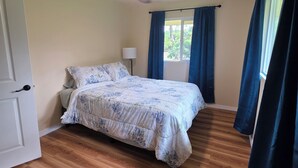 Een slaapkamer met een bed, een plafondventilator, een nachtkastje, een lamp en een raam met gordijnen.