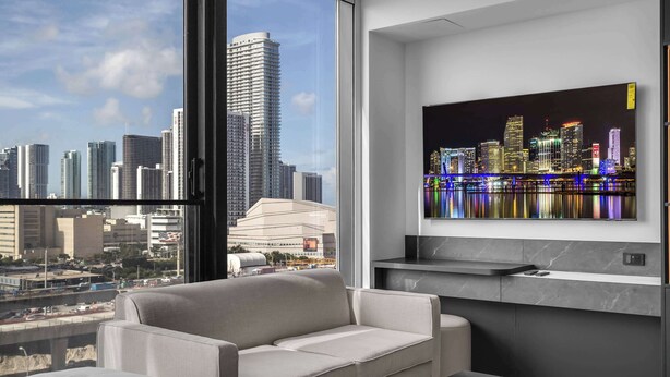 Habitación de hotel moderna con un ventanal grande, un sofá, un televisor montado en la pared y vistas a un horizonte urbano.