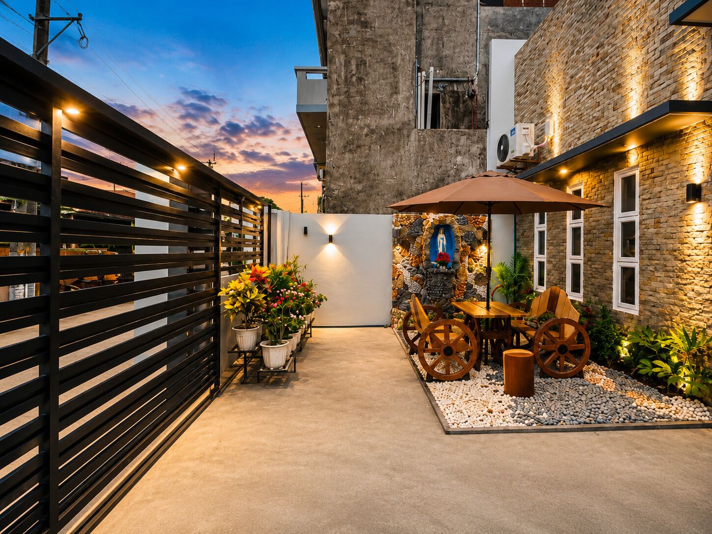 A courtyard with a stone wall, a statue, a wooden table with chairs, and a potted plant.