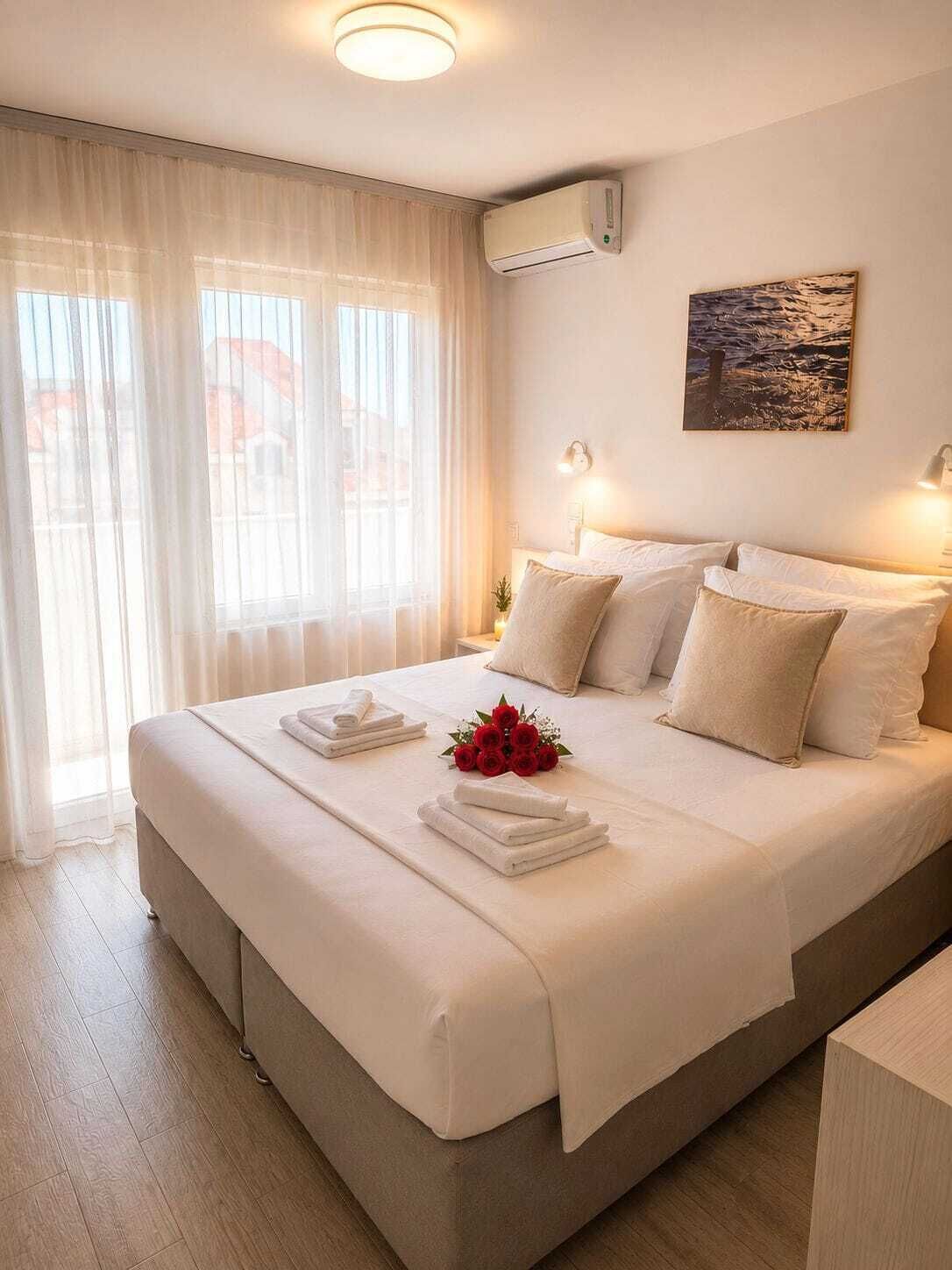 Ett hotellrum med en stor säng, ett sängbord, en luftkonditioneringsanläggning, ett fönster med tunna gardiner och ett konstverk som hänger på väggen.