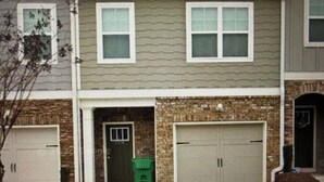 A two-story residential building with a garage, a front door, and a small green trash bin. - Perfect for Nurses-Charming 2-bedroom townhome in Atlanta Metro (Lithonia)