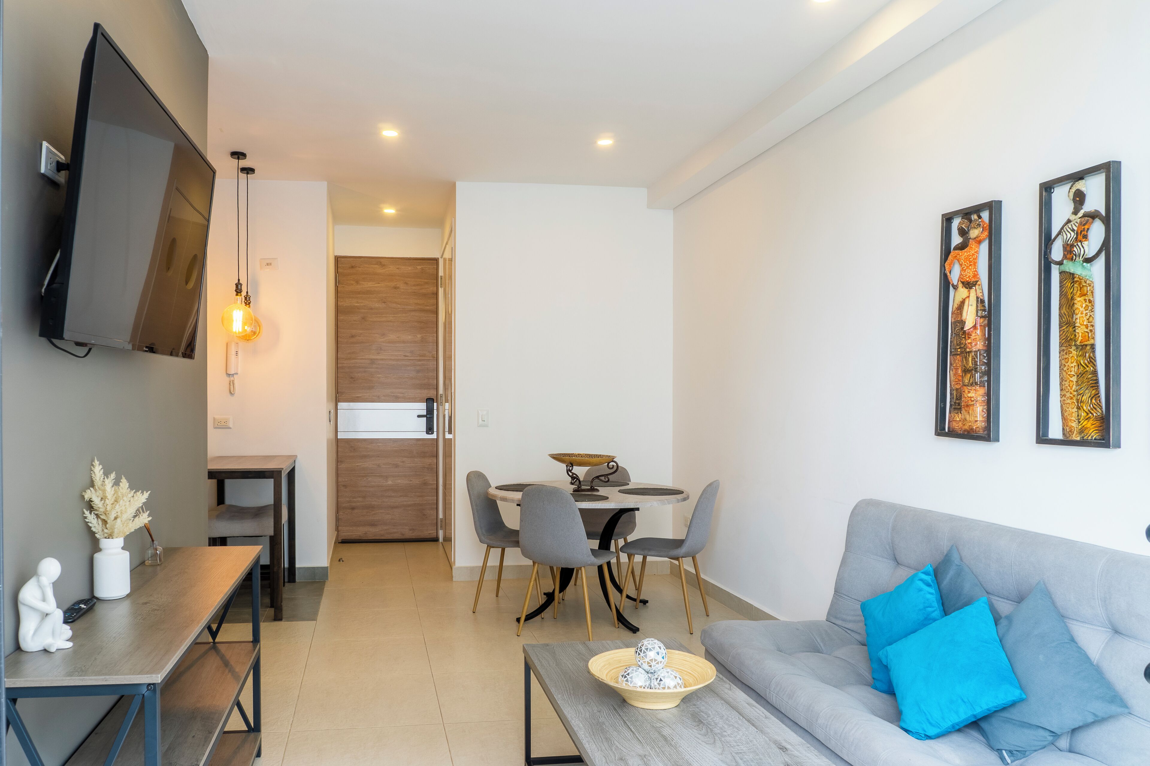 A modern living room with a grey sofa, a dining table with chairs, a TV mounted on the wall, and artwork on the wall.