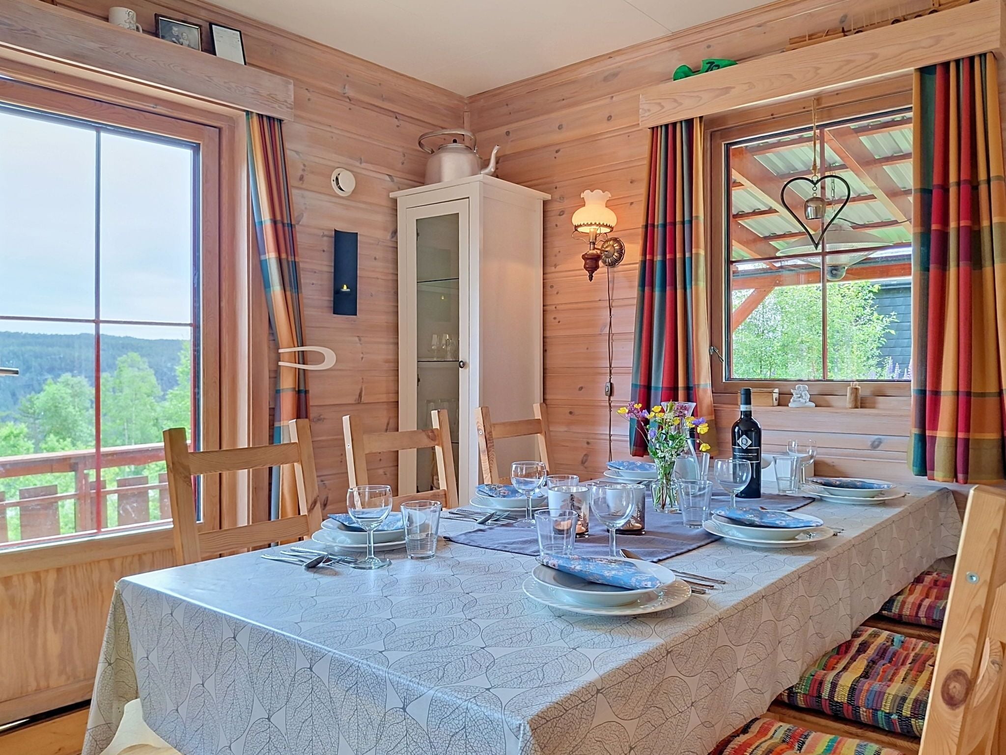 A wooden cabin with a dining table set for four, a window with curtains, and a view of the outdoors.