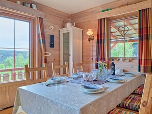 Uma cabana de madeira com uma mesa de jantar posta para quatro pessoas, uma janela com cortinas e vista para o exterior.