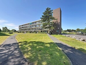 A multi-story building with a flat roof, a large tree in front, and a paved pathway leading to the entrance. - Hyacint with parking by Interhome (De Haan)