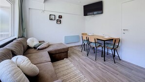 Ein modernes Wohnzimmer mit einer Couch, einem Esstisch mit Stühlen, einem flachbildfernseher an der Wand und einem Fenster mit Vorhängen.