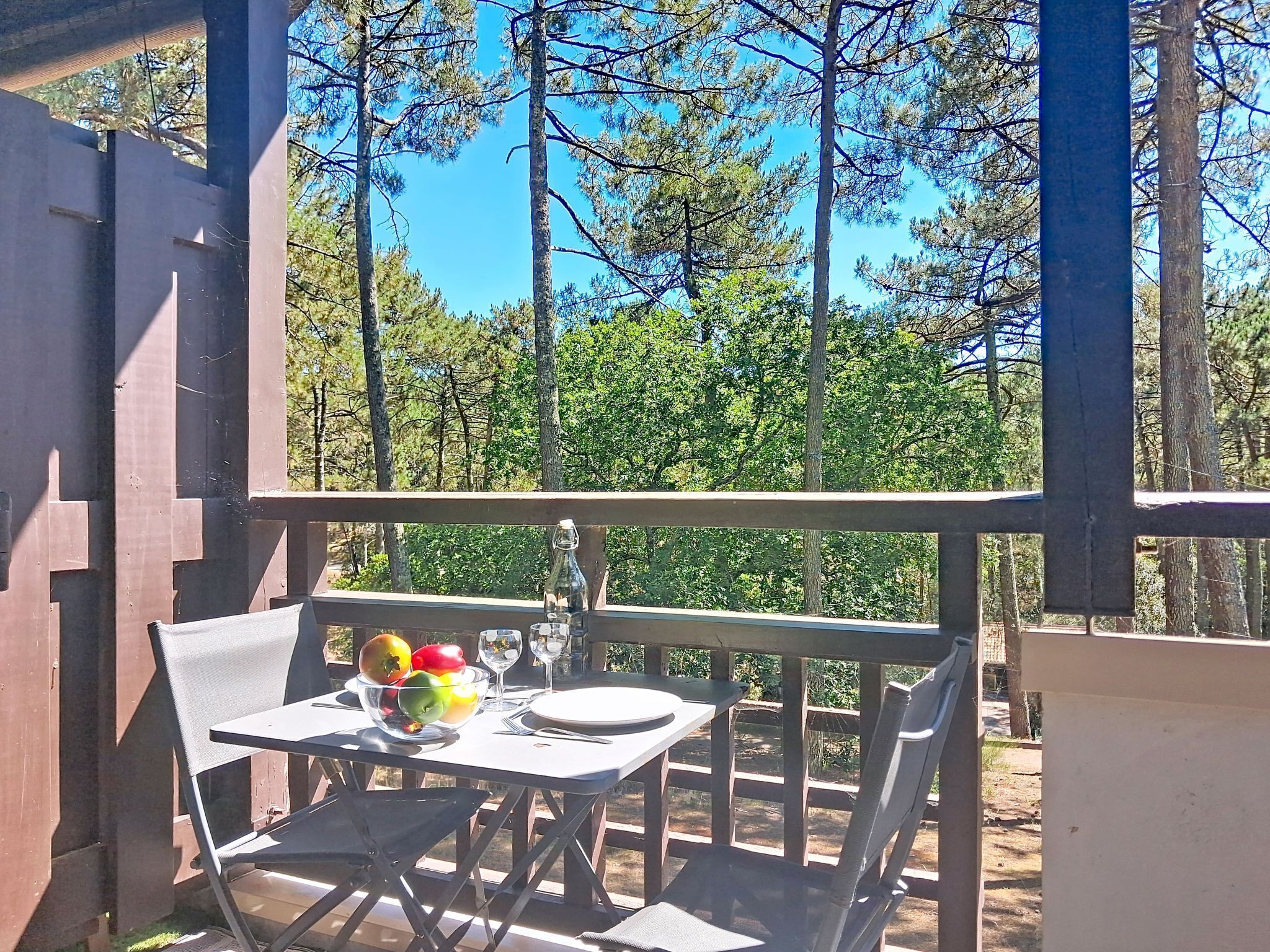 Una mesa con sillas, un bol de fruta y una copa de vino en un balcón con vistas a árboles.