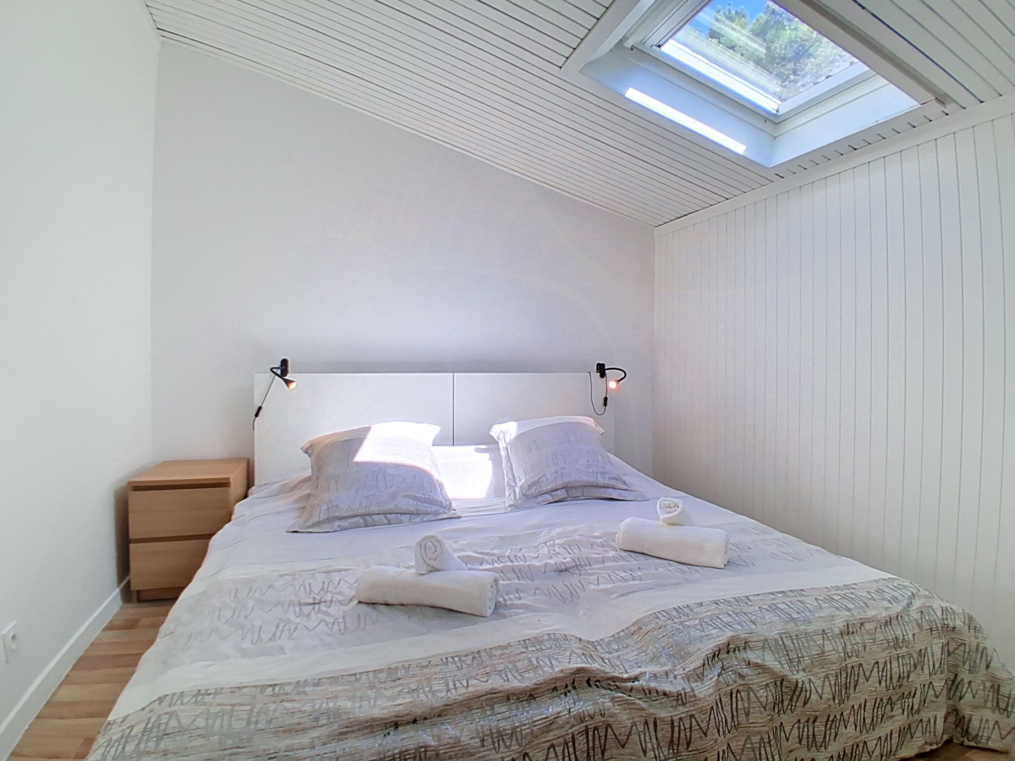 A bedroom with a skylight, a bed with pillows, a nightstand, and a bedside lamp.