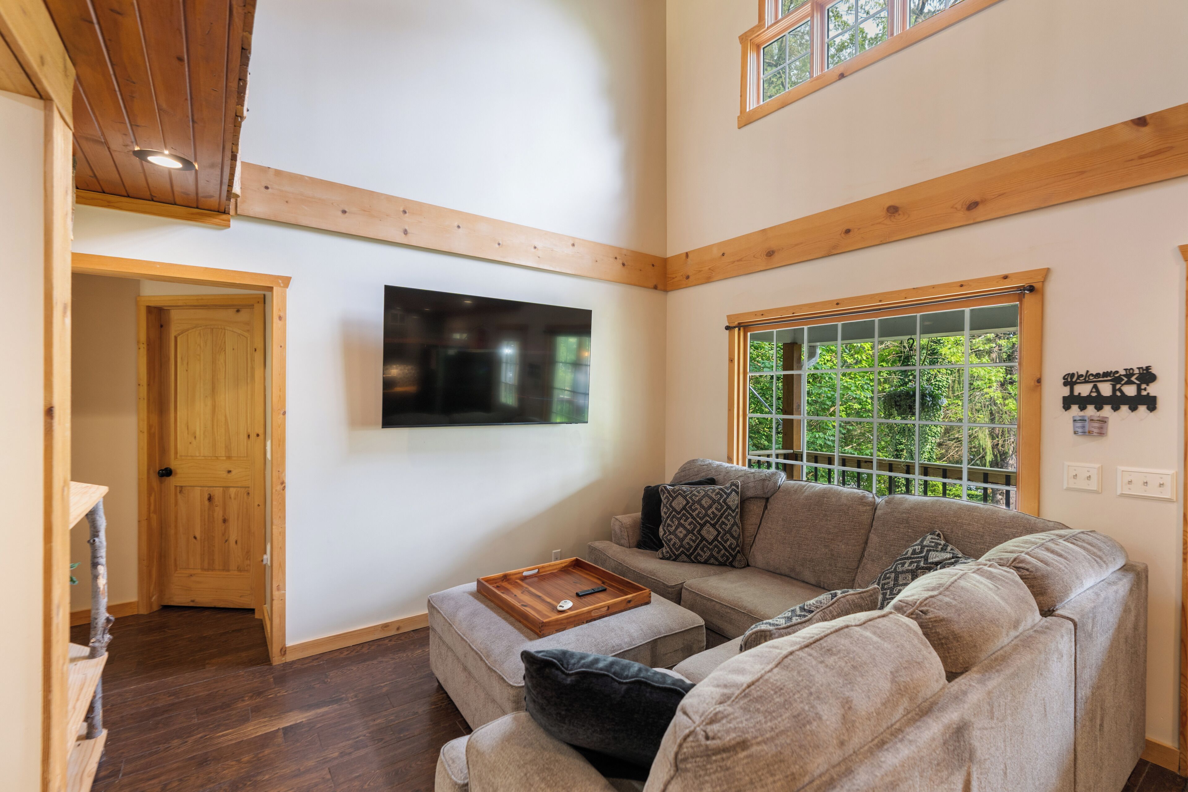 A cozy living room with a large window, a flat-screen TV mounted on the wall, and a wooden ceiling.