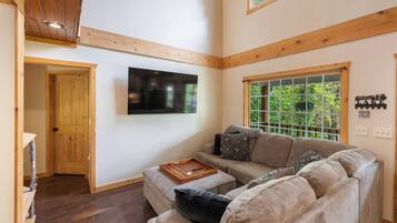 A cozy living room with a large window, a flat-screen TV mounted on the wall, and a wooden ceiling.