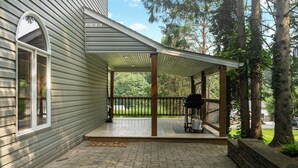 A covered porch with a grill and a propane tank, surrounded by trees and a brick pathway.
