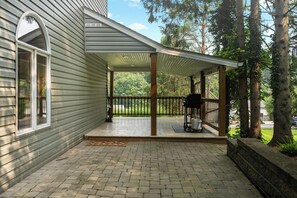 A covered porch with a grill and a propane tank, surrounded by trees and a brick pathway.