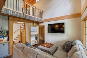 A spacious living area with a high ceiling, a wooden staircase, a flat-screen TV mounted on the wall, and a cozy seating arrangement.