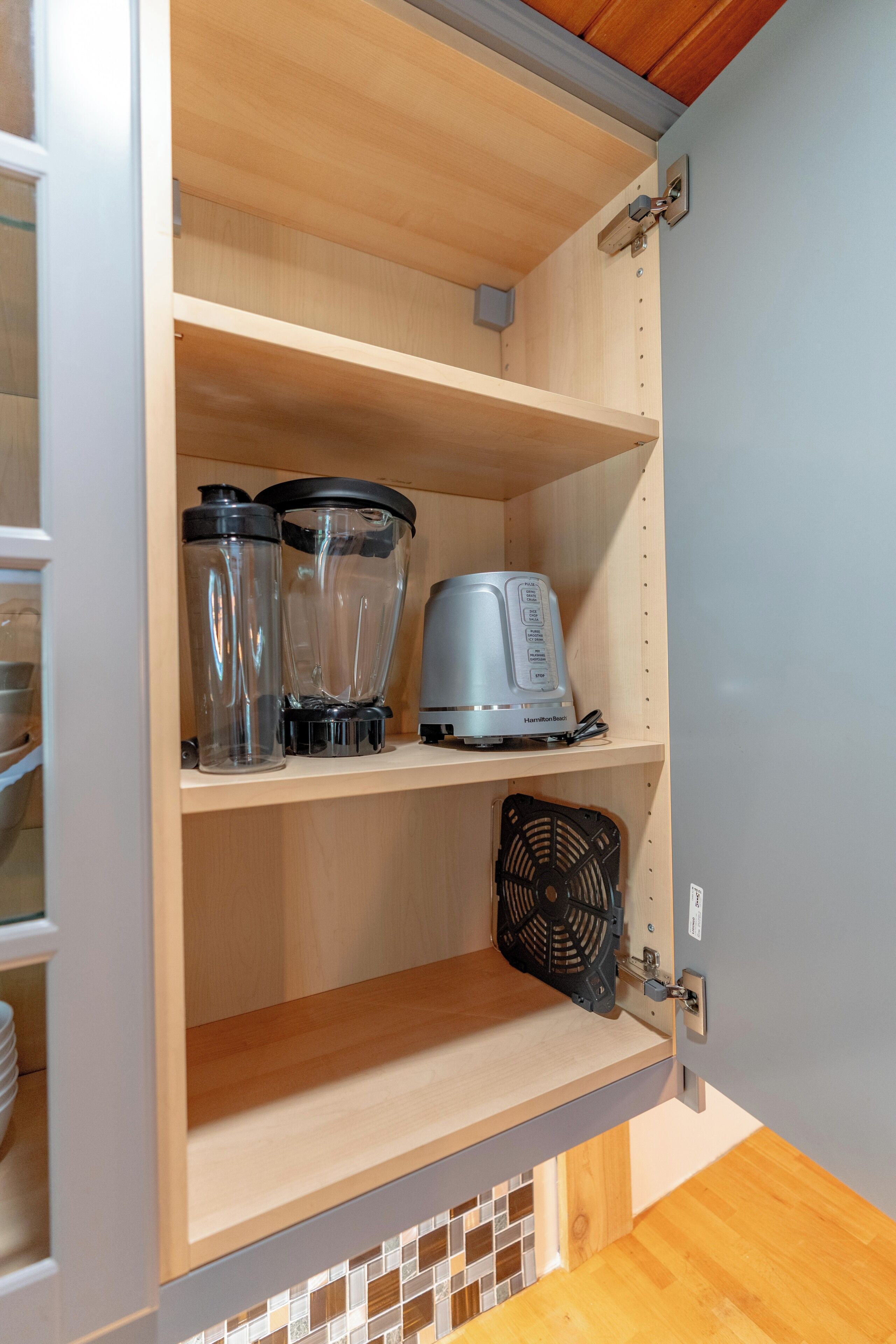 A kitchen cabinet with a blender, a kettle, and a fan on the shelf.