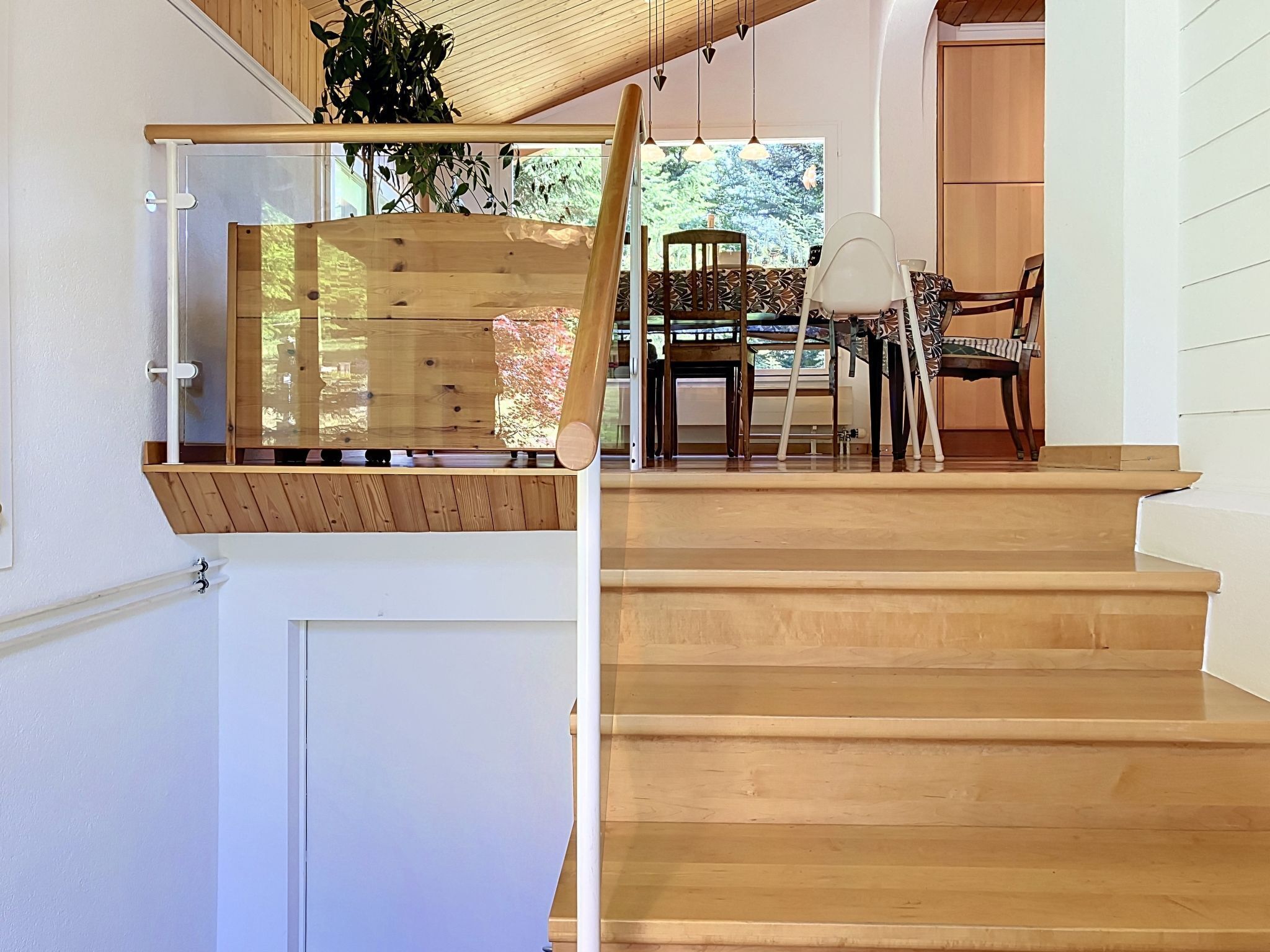 A wooden staircase with a glass railing, a dining area with chairs and a table, and a view of greenery outside.