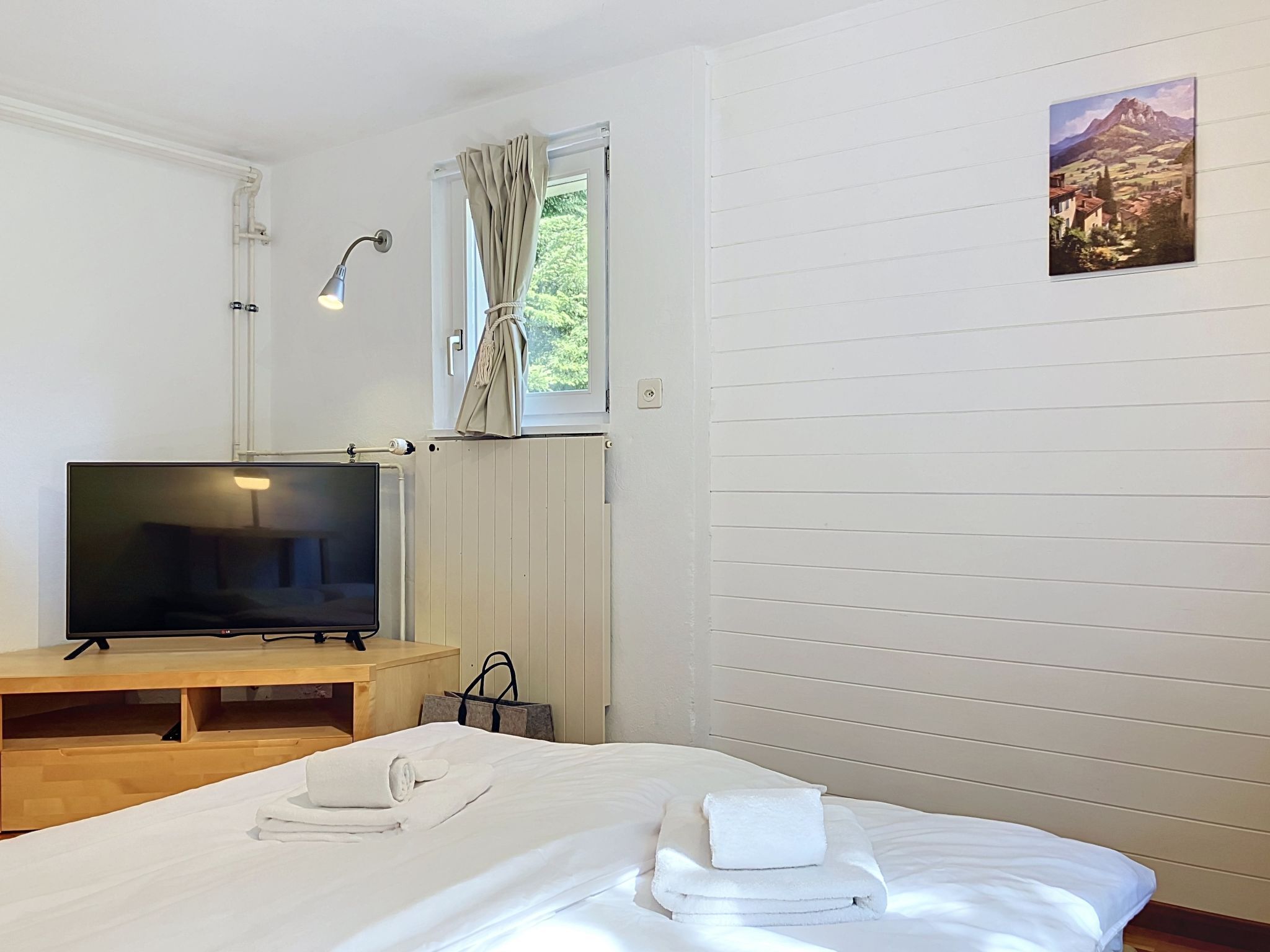 A hotel room with a bed, a TV, a window with curtains, a radiator, and a painting on the wall.