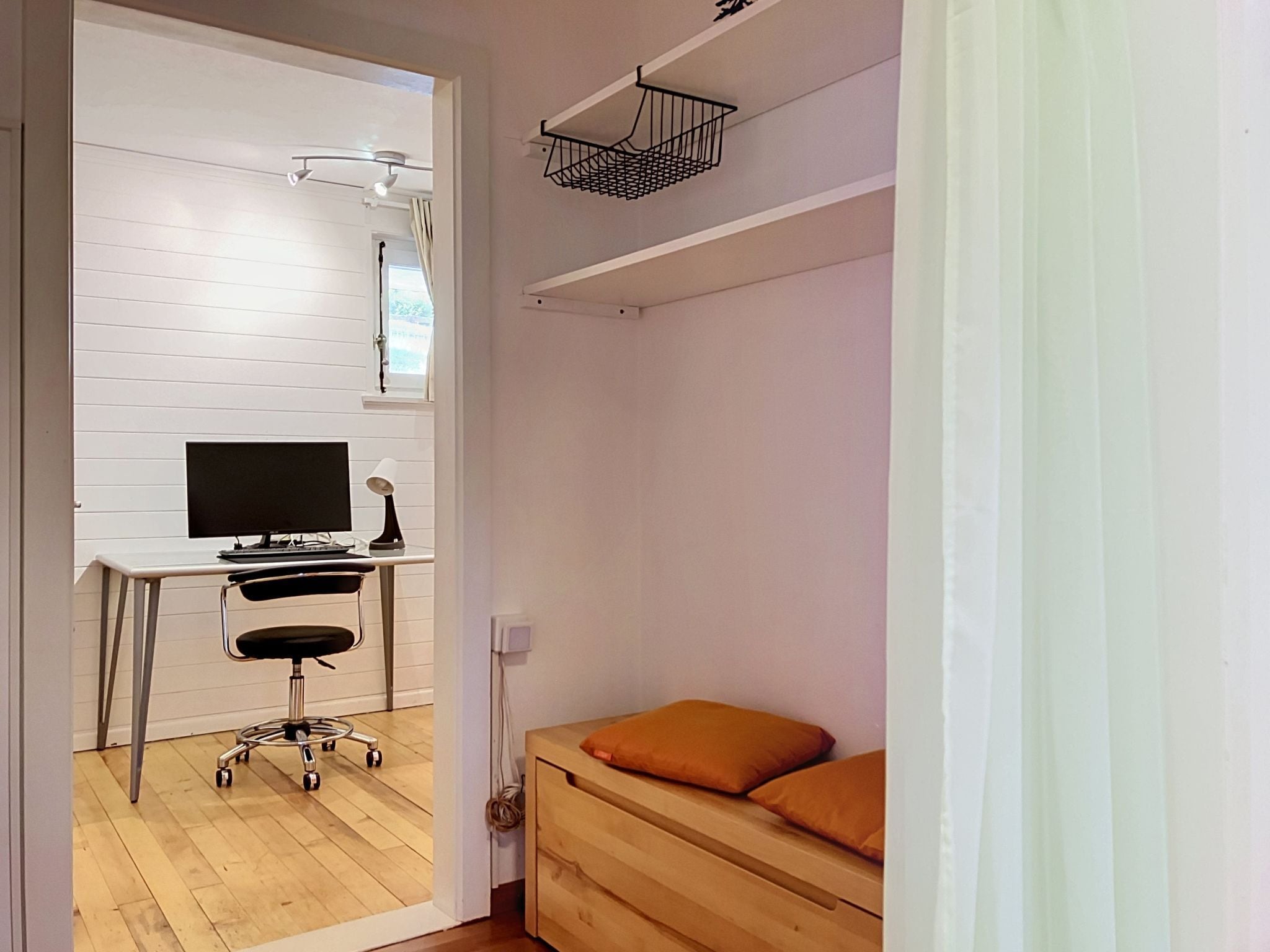 Ein Zimmer mit Schreibtisch, Stuhl, Computer und einem Bett mit orangefarbenem Kissen.