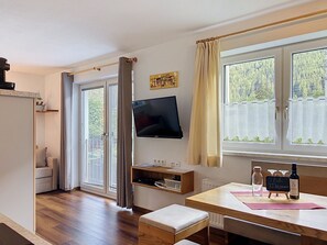 Een kamer met een houten vloer, een bank, een aan de muur bevestigde televisie, een tafel met een fles en glazen, en een raam met gordijnen.