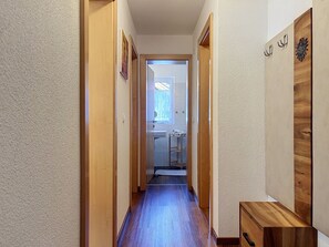 Een smalle gang met houten deuren, een houten bankje en zicht op een kamer met een raam en een deur.