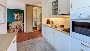 Moderni keittiö, jossa on valkoiset kaapit, sisäänrakennettu uuni sekä ruokailutila, jossa on puiset tuolit ja pöytä.