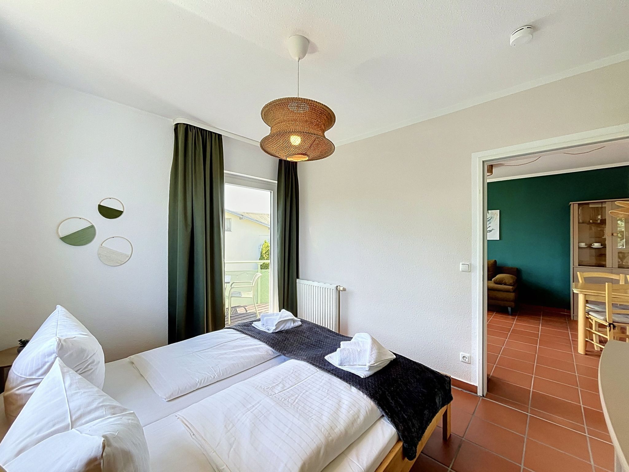 A hotel room with a bed, a hanging light fixture, a view of the outside, and a door leading to a dining area.