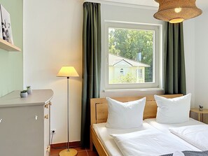 A bedroom with a bed, a bedside table, a lamp, a window with curtains, and a view of a house outside. - Dünenklang by Interhome (Zinnowitz)
