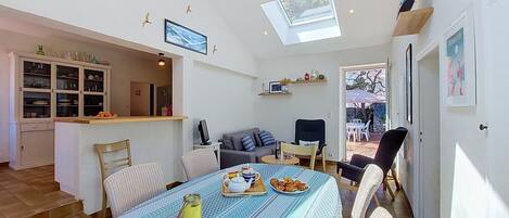 Amplia sala de estar con mesa y sillas de comedor, sofá y televisor. Cuenta con una zona de cocina con armarios y ventana.