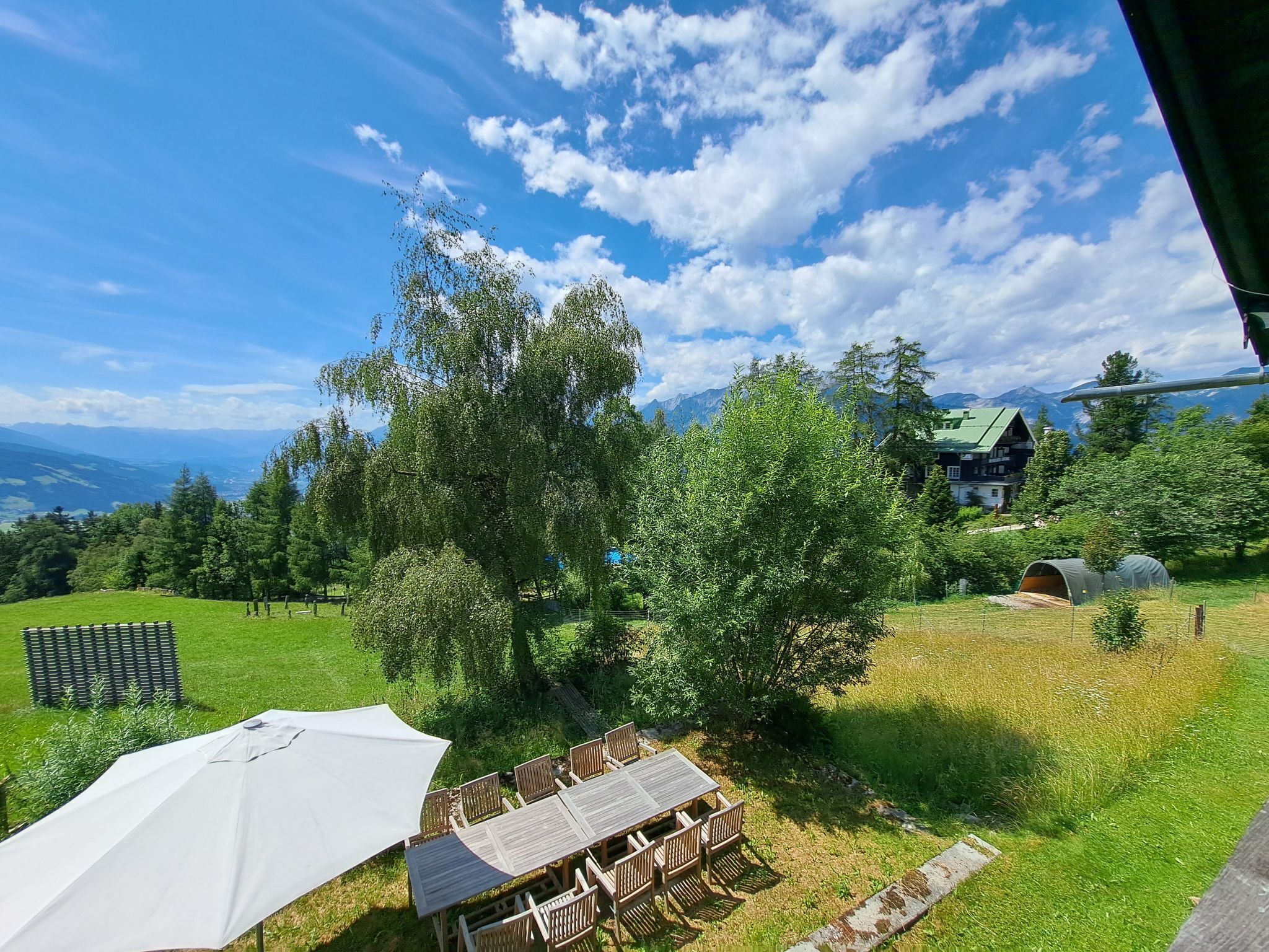 Ein grüner Außenbereich mit Tisch und Stühlen, einem weißen Sonnenschirm und Blick auf Berge im Hintergrund.