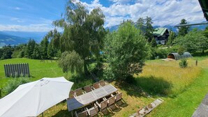 Uma área externa com gramado, uma mesa, cadeiras, um guarda-sol branco e vista para montanhas ao fundo.