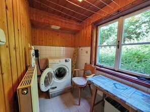 A compact laundry room with a washing machine, a chair, and a window with a view of greenery.