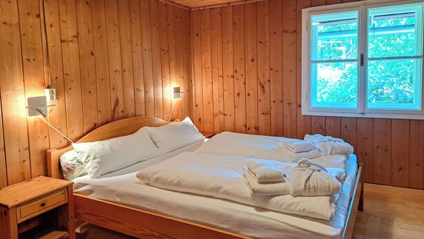 Quarto com painéis de madeira, uma cama, mesa de cabeceira e janela com vista para o verde.