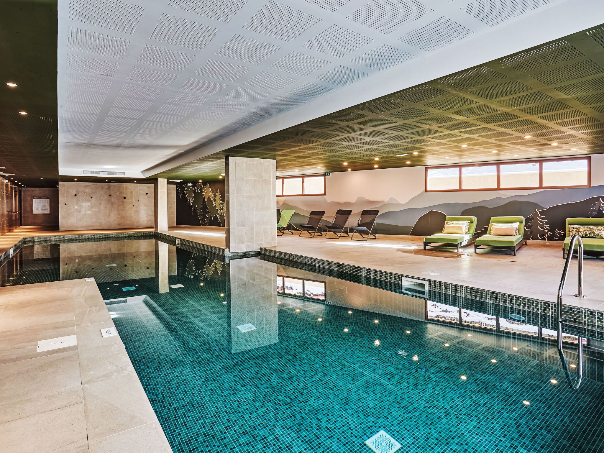 Una piscina interior con suelo de baldosas, sillones de descanso y un mural en la pared.
