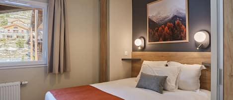 A hotel room with a bed, a window with a view of buildings, a painting on the wall, and two wall-mounted lights.