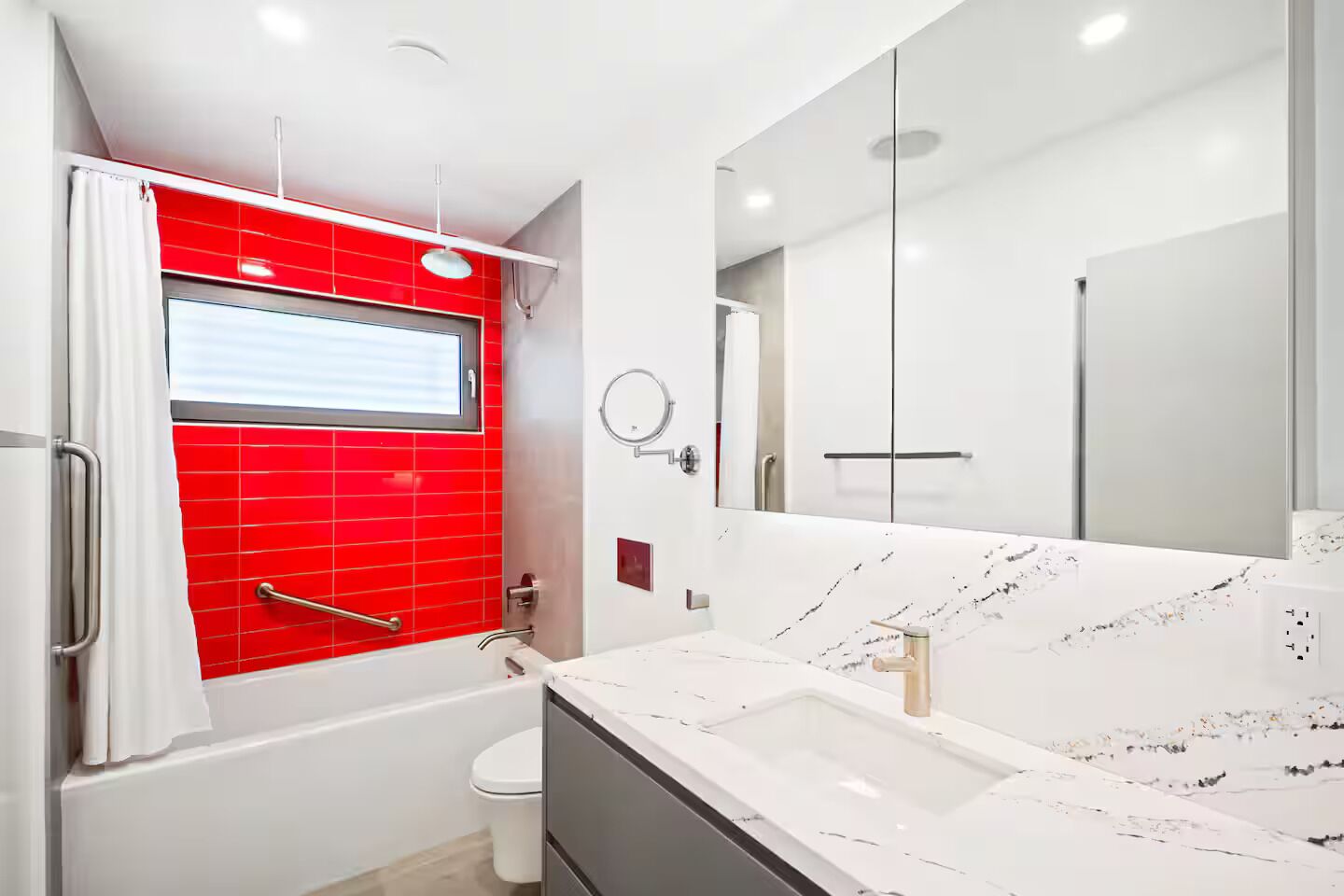 A modern bathroom with a red-tiled shower enclosure, a white sink with a marble countertop, and a large mirror.