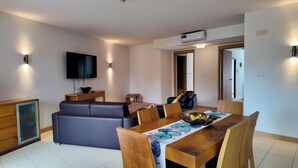 En moderne stue med sofa, fjernsyn, spisebord med stole og et køkkenområde.