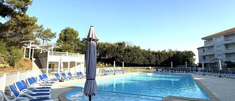 Una piscina con sillas de descanso y sombrillas, rodeada de árboles y un edificio.