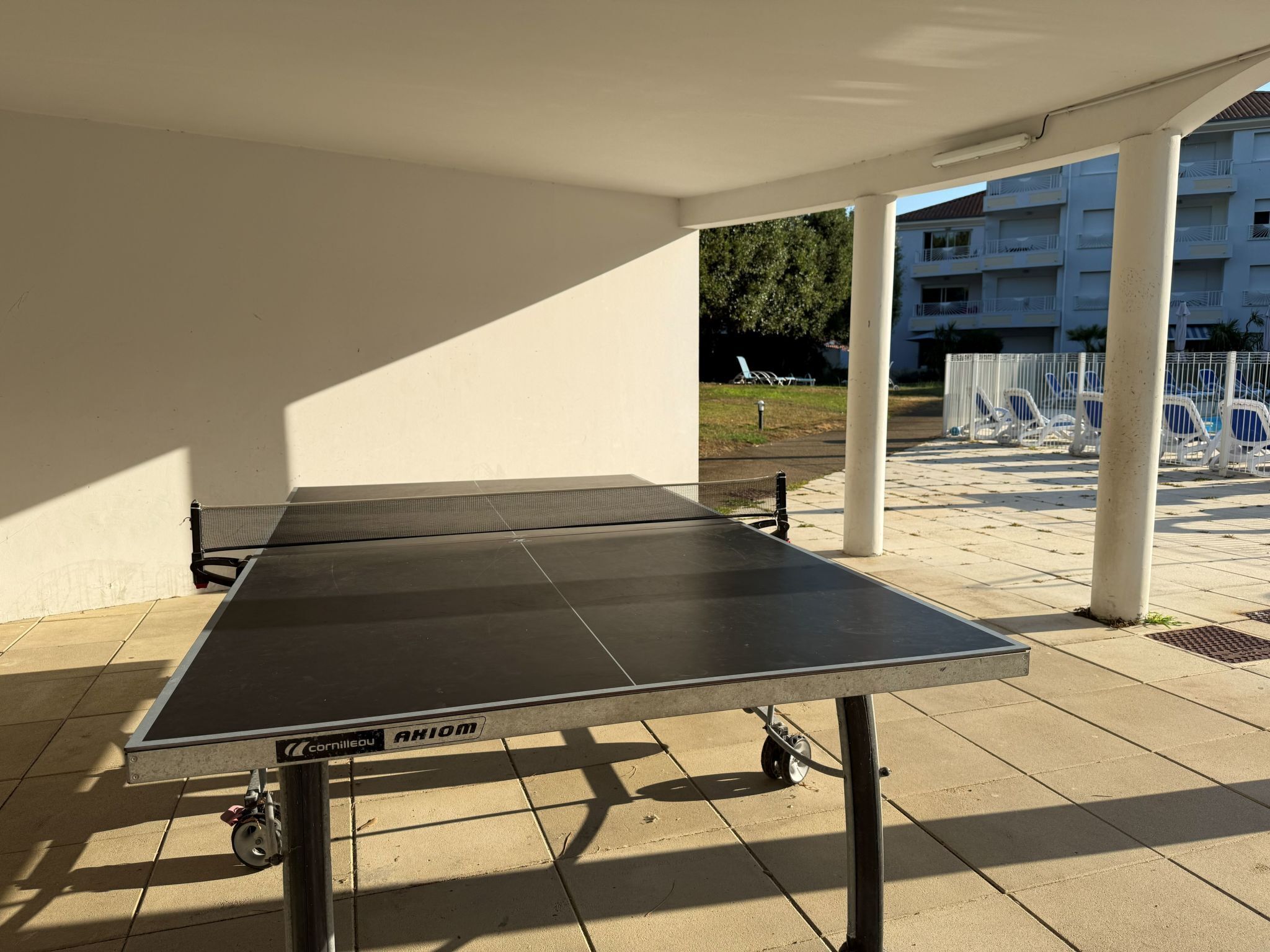 Une table de ping-pong installée à l’extérieur, avec un filet, sur une terrasse carrelée, avec un bâtiment blanc en arrière-plan.