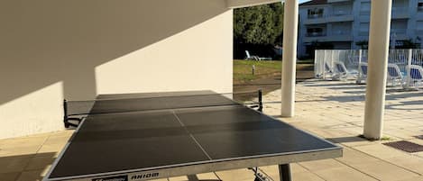 Ein Tischtennis-Tisch mit Netz steht auf einer gefliesten Terrasse, im Hintergrund ist ein weißes Gebäude zu sehen.