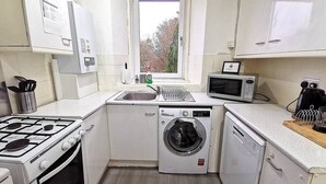 Een compacte keuken met gasfornuis, wasmachine, magnetron, en een raam met uitzicht op bomen.