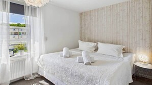 Une chambre d’hôtel avec un grand lit, une fenêtre ornée de rideaux légers, une plante en pot et un luminaire décoratif.
