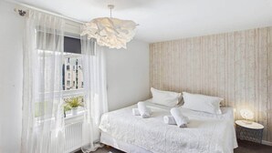 Une chambre à coucher avec un lit, une fenêtre avec des rideaux légers, un luminaire suspendu et une table de chevet avec une lampe.