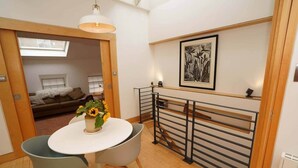 Een compacte kamer met een ronde tafel, twee stoelen, een potplant, een plafondlamp, een lijstwerk en uitzicht op een andere kamer met een bed en een raam.