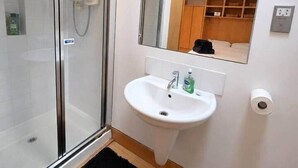 Een badkamer met een douchecabine van glas, een witte wastafel, een spiegel en een zwart vloerkleed.