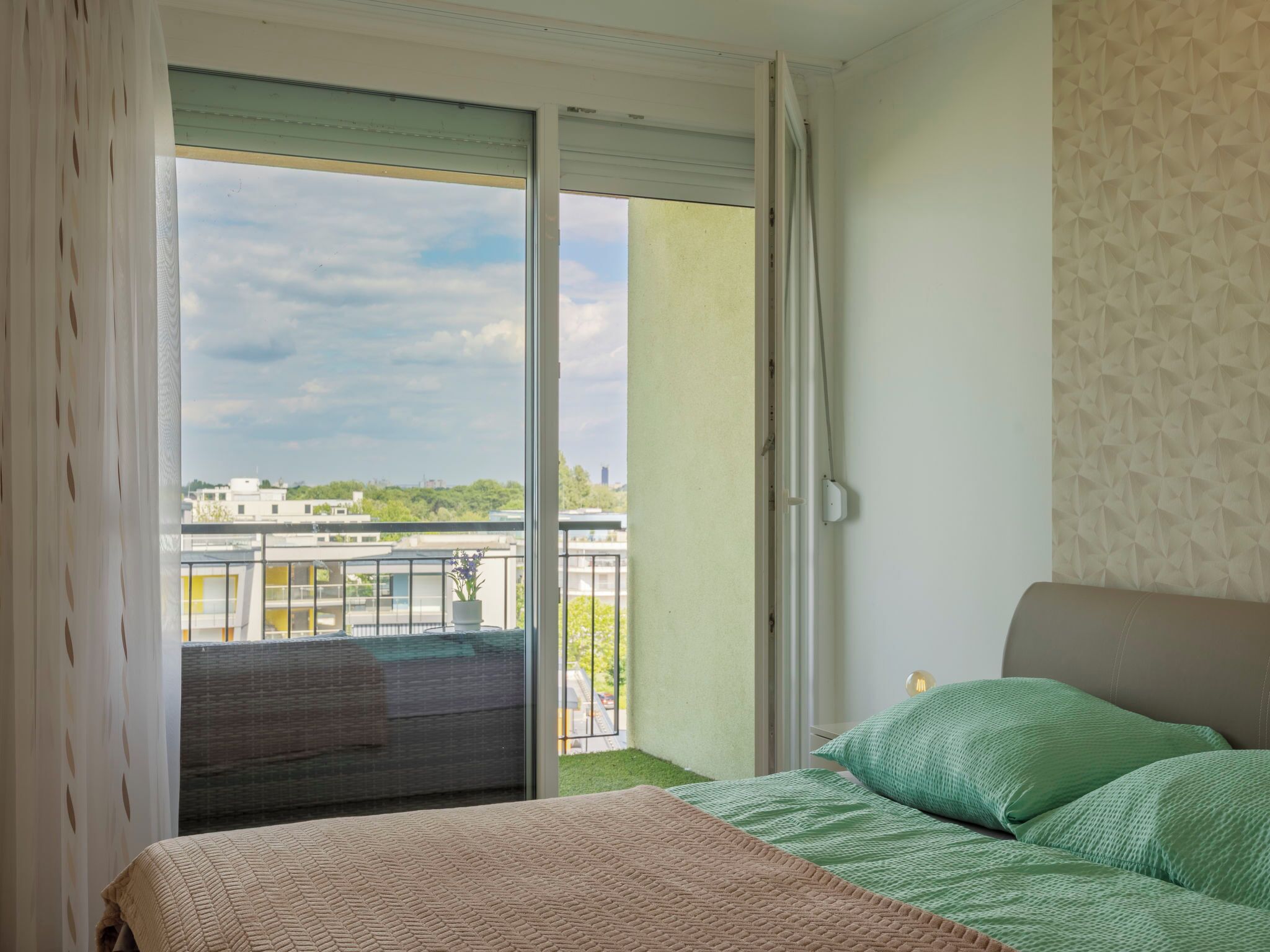 Een slaapkamer met een bed, groene kussens, een raam met uitzicht op gebouwen en een balkon.