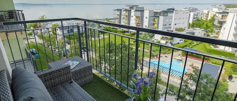Un balcón con un sofá, una mesita con una planta en maceta y vistas a una piscina y edificios.