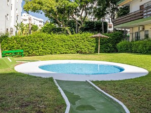 A swimming pool with a surrounding grassy area, a bench, and a thatched roof structure. - Las Naciones by Interhome (Benalmádena Costa)