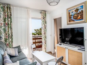A living room with a sofa, a TV, a small table, and a balcony with a view of buildings and trees. - Las Naciones by Interhome (Benalmádena Costa)